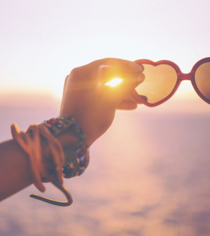 Hand holding heart-shaped sunglasses on a beach at sunset